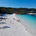 A group of people walk and relax on a white sandy beach beside clear blue water, with forested land in the background under a clear sky.