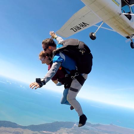 Two people tandem skydiving, free-falling in midair with parachute packs, as a small airplane flies away above coastline and ocean.