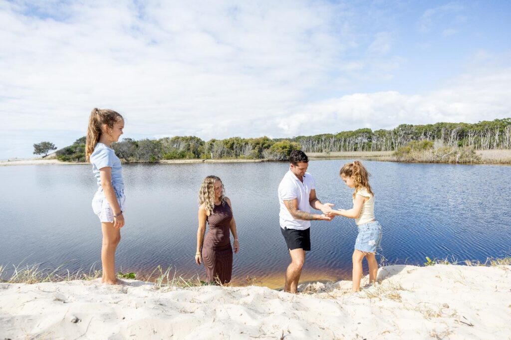 Four people stand on sandy ground by a lake; one man helps a young girl up a small slope, while another woman and girl watch. Trees and partly cloudy sky are in the background.