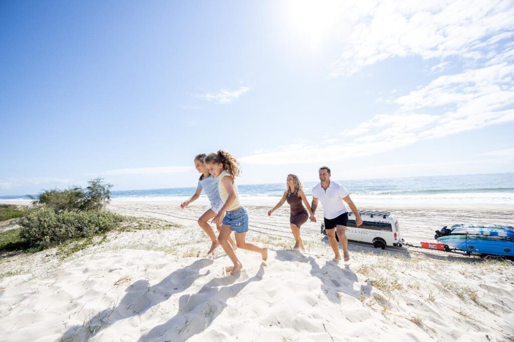 Four people walk up a sandy dune at the beach on a sunny day, with a van and surfboards parked near the shoreline in the background.