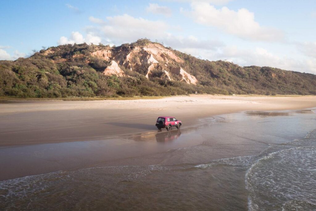 A red SUV drives along a wide, sandy beach with a rocky hill and greenery in the background under a partly cloudy sky.