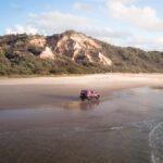 A red SUV drives along a wide, sandy beach with a rocky hill and greenery in the background under a partly cloudy sky.