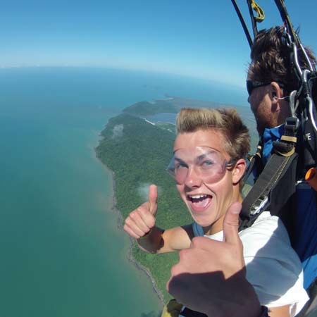 Two people are tandem skydiving above a coastline; one is giving a double thumbs-up and smiling, wearing clear protective goggles.