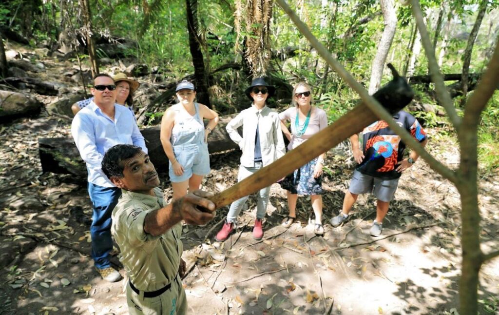 A guide demonstrates a tool to a group of six people in an outdoor forest setting during daylight.