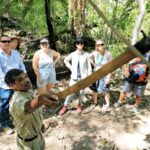 A guide demonstrates a tool to a group of six people in an outdoor forest setting during daylight.