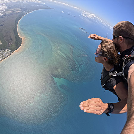 Two people tandem skydiving above a coastline with clear blue water, sandy beaches, and scattered clouds visible in the distance.