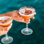 Two hands hold cocktail glasses with foam, a dried citrus slice, and chocolate-rimmed edges, against a blurred blue water background.