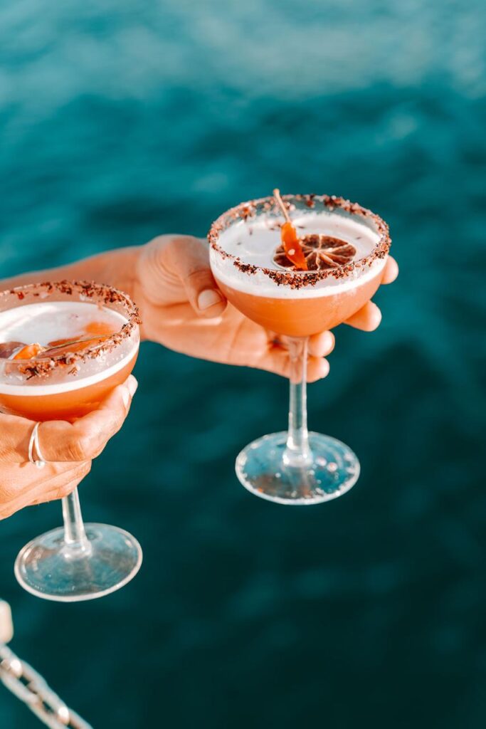 Two hands hold cocktail glasses with foam, a dried citrus slice, and chocolate-rimmed edges, against a blurred blue water background.