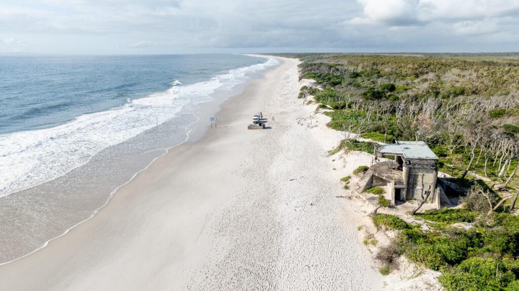 Aerial view of a long, sandy beach with ocean waves on the left, vegetation on the right, and an old concrete structure in the foreground near the shore.