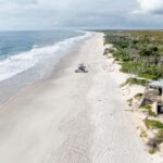 Aerial view of a long, sandy beach with ocean waves on the left, vegetation on the right, and an old concrete structure in the foreground near the shore.