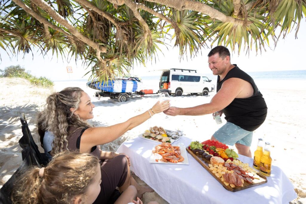 A man and woman sit at a table with seafood and fruit on a beach, exchanging drinks. A van and kayak are parked nearby under a tree, with the ocean in the background.