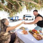 A man and woman sit at a table with seafood and fruit on a beach, exchanging drinks. A van and kayak are parked nearby under a tree, with the ocean in the background.