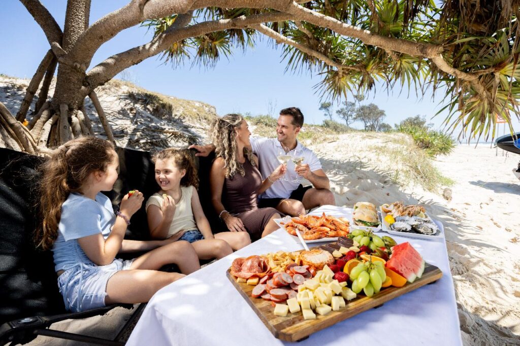 A family of four sits under a tree on a sandy beach, enjoying a picnic with assorted fruits, cheeses, meats, and seafood on a table.