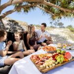 A family of four sits under a tree on a sandy beach, enjoying a picnic with assorted fruits, cheeses, meats, and seafood on a table.