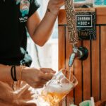 A person pours beer from a tap labeled "JARAH BOY" into a glass mug at an outdoor bar setup.