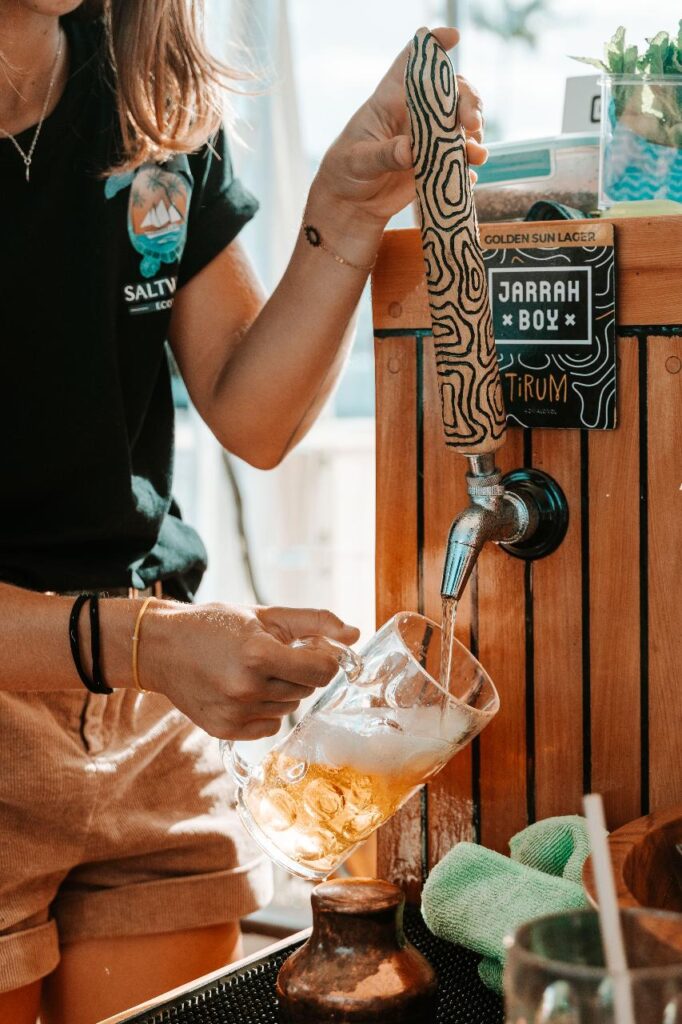 A person pours beer from a tap labeled "JARAH BOY" into a glass mug at an outdoor bar setup.