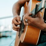 Close-up of a person playing an acoustic guitar outdoors, showing hands with rings and tattoos, with a blurred marina background.