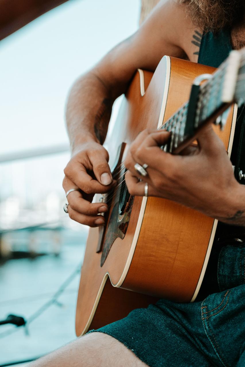Close-up of a person playing an acoustic guitar outdoors, showing hands with rings and tattoos, with a blurred marina background.