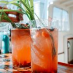 Two tall glasses of iced orange-pink drink with fruit and green garnish sit on a wooden table aboard a boat in sunlight.