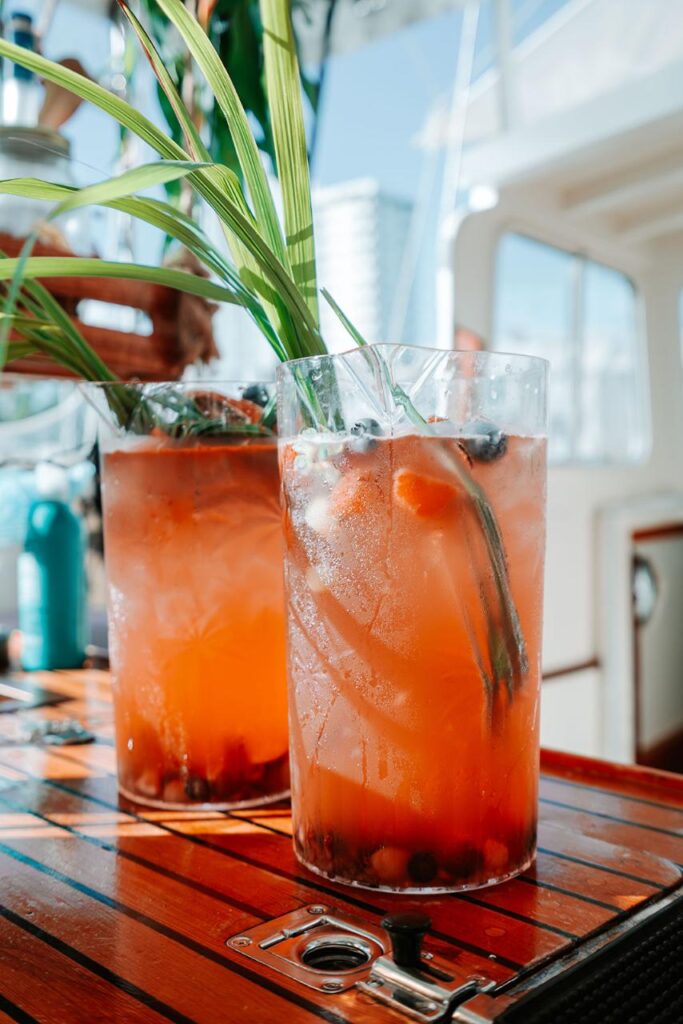 Two tall glasses of iced orange-pink drink with fruit and green garnish sit on a wooden table aboard a boat in sunlight.