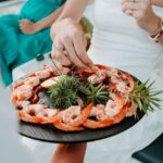 A person in a white dress takes shrimp from a black platter garnished with greens and lemon slices. Another person in a green dress is seated in the background.