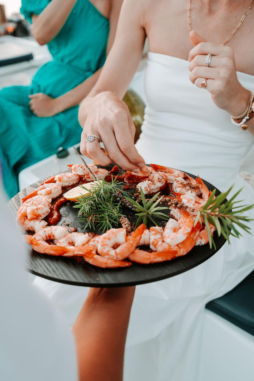 A person in a white dress takes shrimp from a black platter garnished with greens and lemon slices. Another person in a green dress is seated in the background.
