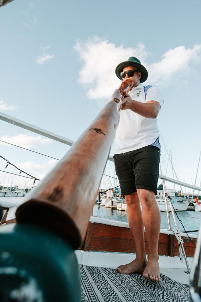 A person standing barefoot on a boat deck plays a long wooden wind instrument with marina and sailboats in the background.