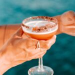 A person holding a cocktail in a coupe glass with a chocolate rim, against a blurred blue background.