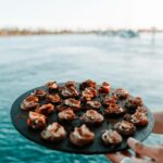 A person holds a round black tray filled with bite-sized appetizers near a body of water, with a blurred shoreline in the background.