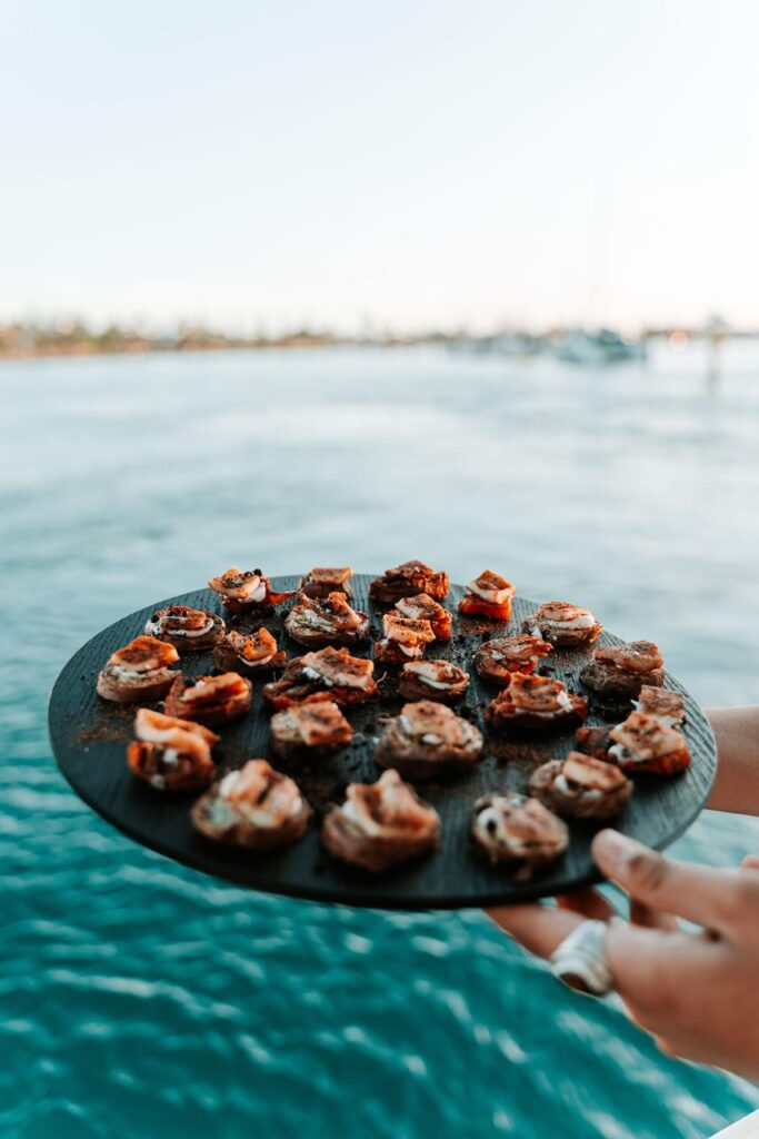 A person holds a round black tray filled with bite-sized appetizers near a body of water, with a blurred shoreline in the background.