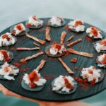 A round black tray holds several small appetizers, each featuring a cracker topped with white cream and a reddish garnish, arranged in a circular pattern.
