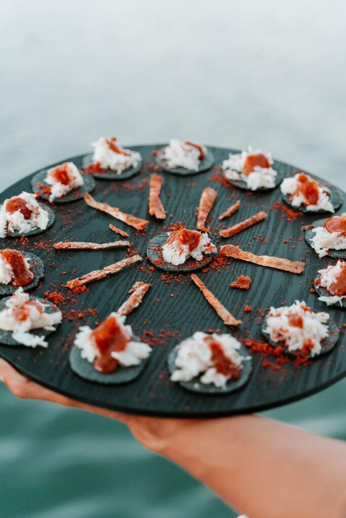 A round black tray holds several small appetizers, each featuring a cracker topped with white cream and a reddish garnish, arranged in a circular pattern.