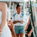 A woman in a white shirt and denim shorts holds a tray of appetizers while standing among people on a boat.