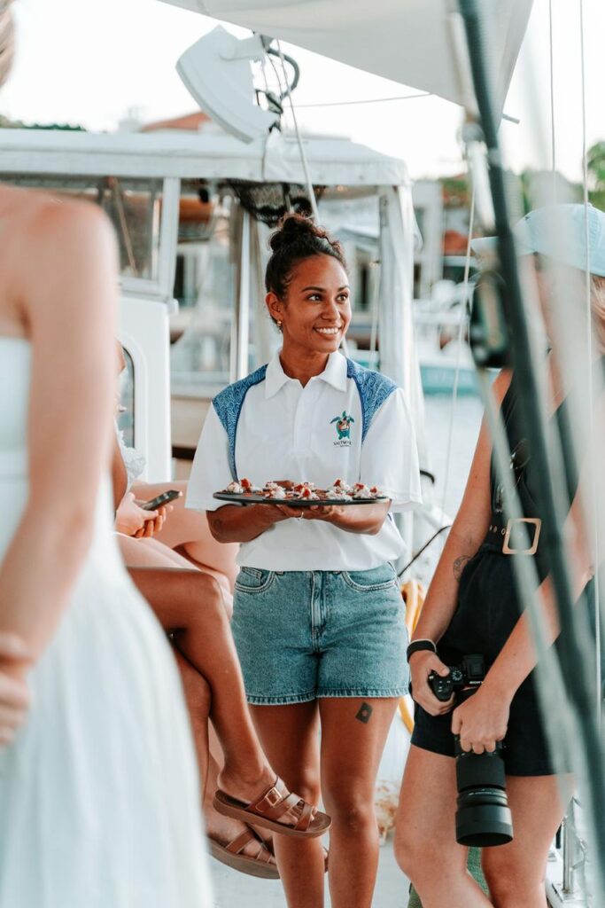 A woman in a white shirt and denim shorts holds a tray of appetizers while standing among people on a boat.