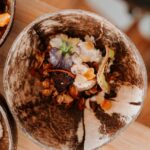 A close-up of a rustic bowl containing granola, dried fruit, and edible flowers, placed on a wooden surface.