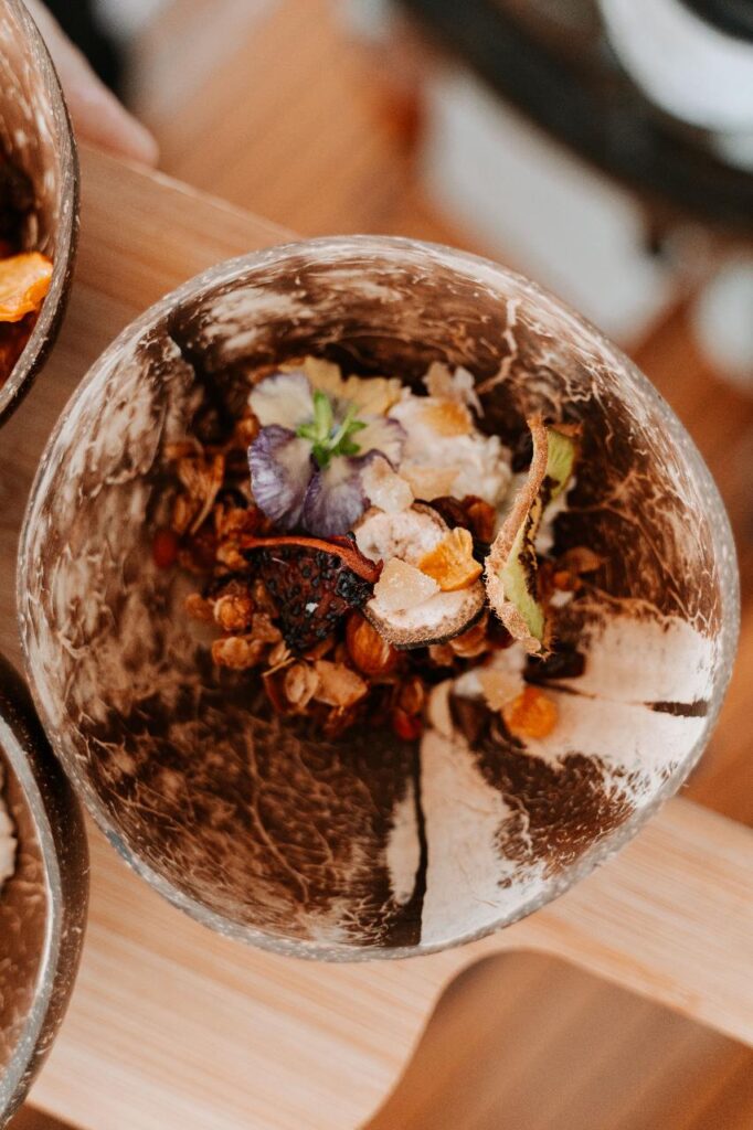 A close-up of a rustic bowl containing granola, dried fruit, and edible flowers, placed on a wooden surface.