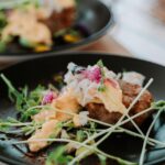 A black plate with a gourmet dish featuring a poached egg, microgreens, sauce, and a round piece of meat or patty, with another similar plate blurred in the background.