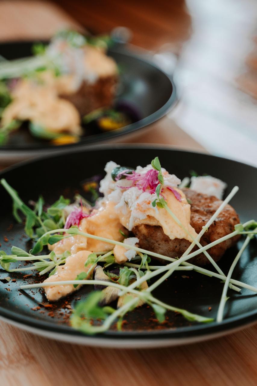 A black plate with a gourmet dish featuring a poached egg, microgreens, sauce, and a round piece of meat or patty, with another similar plate blurred in the background.