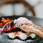 A plated dish featuring a piece of glazed meat on a waffle, garnished with a dried lotus root slice, cream, and herbs, lightly dusted with powdered sugar.