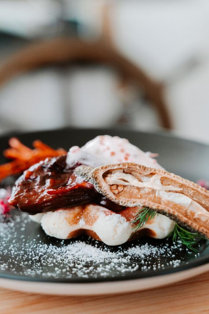 A plated dish featuring a piece of glazed meat on a waffle, garnished with a dried lotus root slice, cream, and herbs, lightly dusted with powdered sugar.