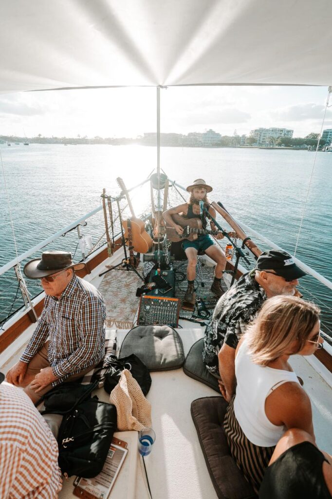 A musician plays guitar on the deck of a boat while several people sit and relax around him; water and buildings are visible in the background.