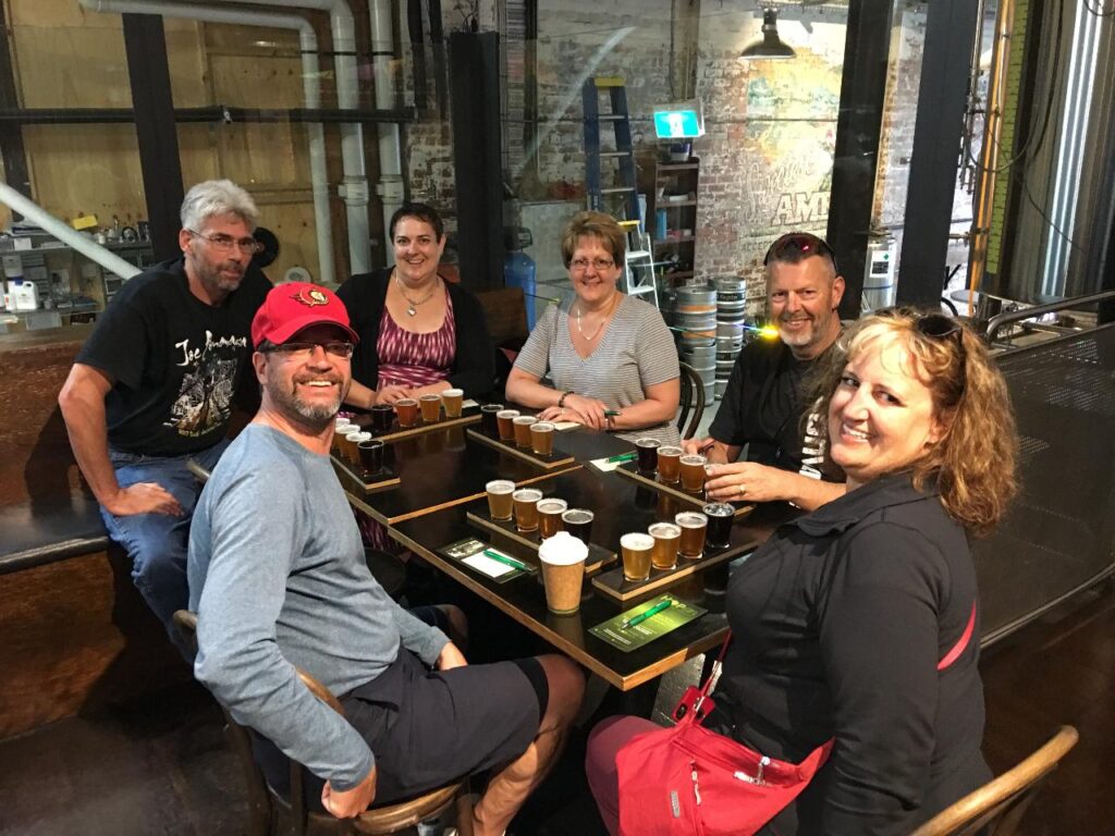 Six adults sit around a table with several beer flights and drinks in a rustic indoor brewery setting, smiling at the camera.