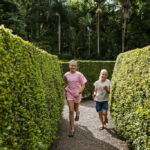 Two children run along a gravel path between tall, neatly trimmed green hedges in an outdoor garden or maze setting.
