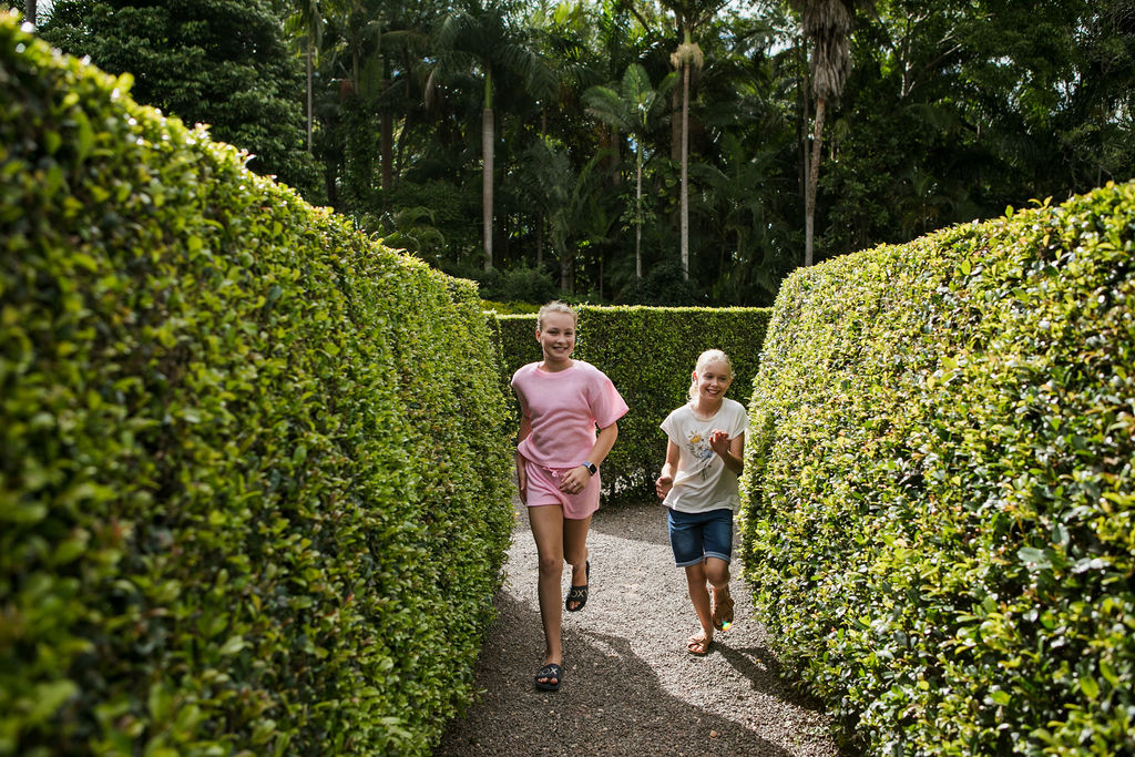 Two children run along a gravel path between tall, neatly trimmed green hedges in an outdoor garden or maze setting.