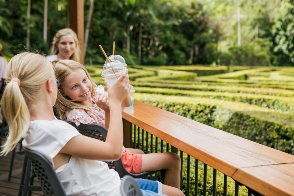 Two girls sitting at an outdoor wooden railing, holding plastic cups with straws, with a maze and greenery in the background. An adult woman sits behind them.