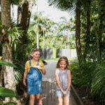Two young children walk along a wooden path surrounded by lush green tropical plants and trees, smiling and enjoying the outdoors.