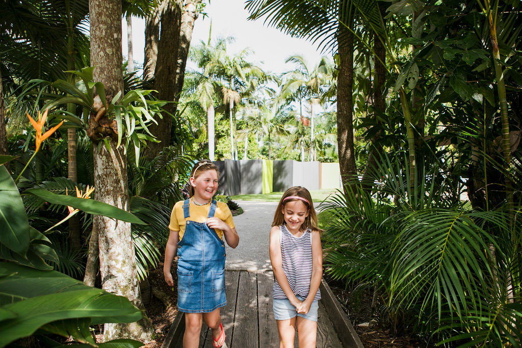 Two young children walk along a wooden path surrounded by lush green tropical plants and trees, smiling and enjoying the outdoors.