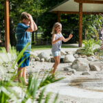 Three children play barefoot near a small water fountain outdoors, with one child touching the spray and another running nearby. Trees and a covered area are in the background.