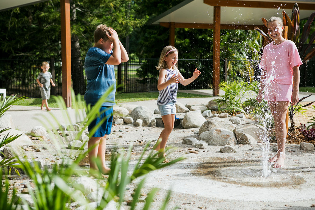 Three children play barefoot near a small water fountain outdoors, with one child touching the spray and another running nearby. Trees and a covered area are in the background.