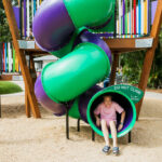 A girl wearing a pink shirt exits a spiral playground slide while another girl walks nearby on the playground's sandy surface.